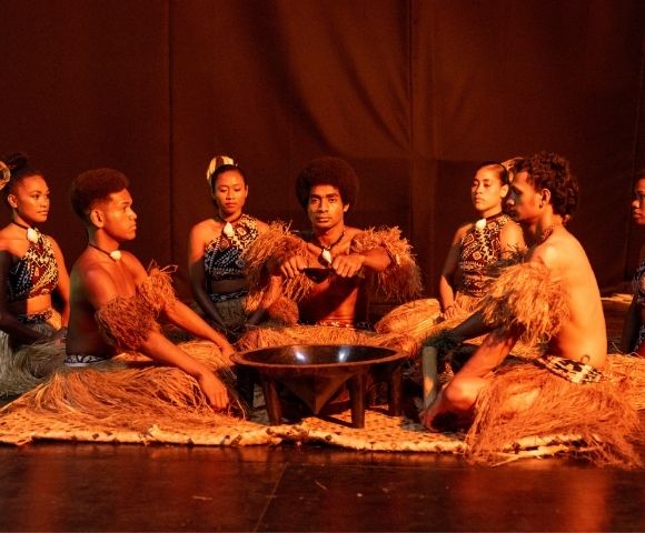 A group of performers in traditional attire sitting in a circle around a small fire on stage.
