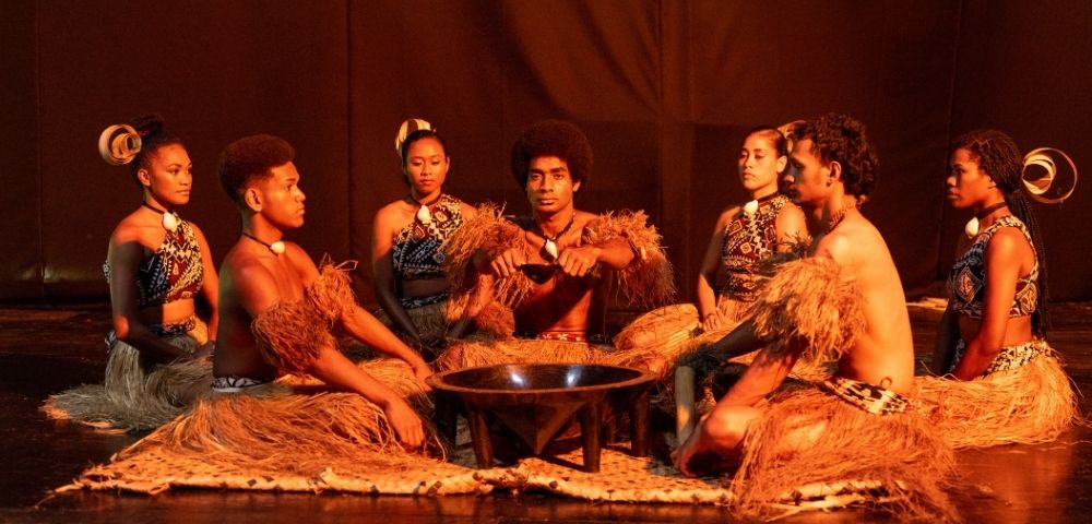 A group of performers in traditional attire sitting in a circle around a small fire on stage.