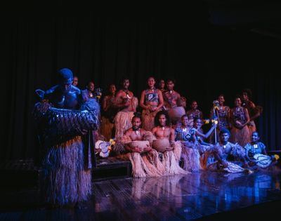 A group of performers in traditional attire, featuring grass skirts and body paint, pose on stage with various musical instruments.