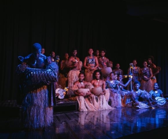 A group of performers in traditional attire, featuring grass skirts and body paint, pose on stage with various musical instruments.