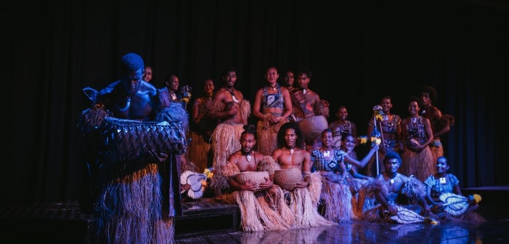 A group of performers in traditional attire, featuring grass skirts and body paint, pose on stage with various musical instruments.