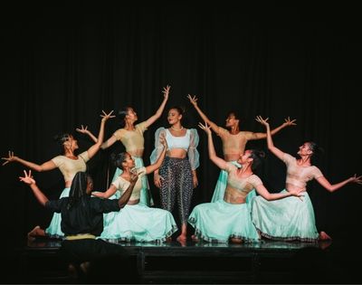 A group of dancers on stage, some in traditional attire, performing with expressive poses against a dark background.