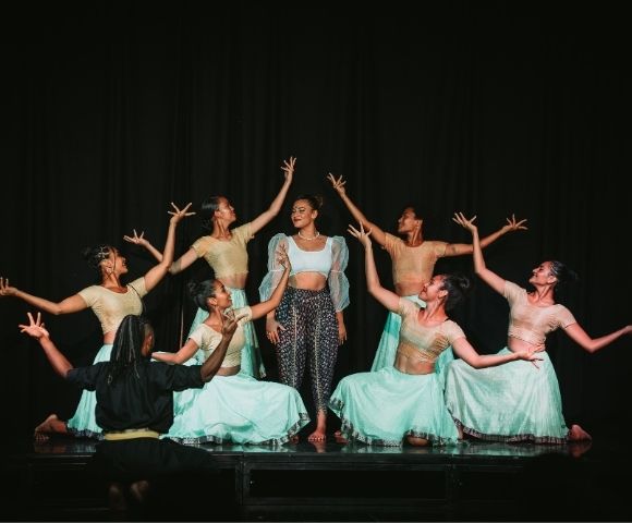 A group of dancers on stage, some in traditional attire, performing with expressive poses against a dark background.