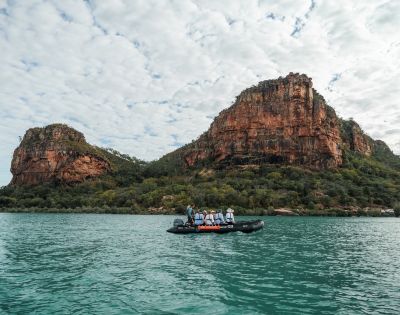 A group of people in a small boat on calm water, surrounded by rocky cliffs and a cloudy sky.