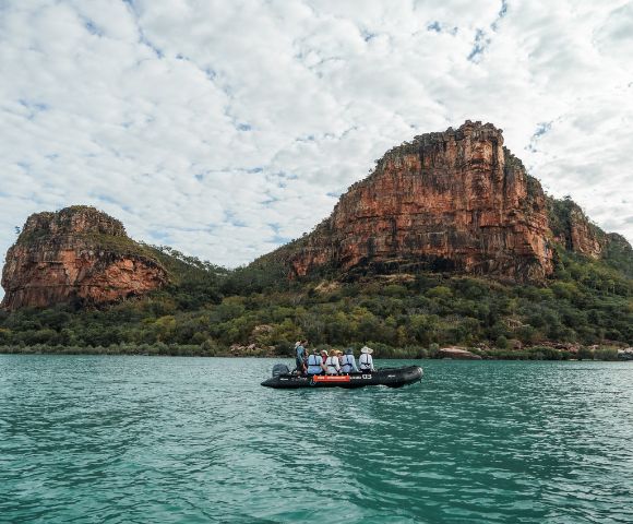 A group of people in a small boat on calm water, surrounded by rocky cliffs and a cloudy sky.