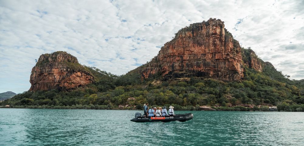 A group of people in a small boat on calm water, surrounded by rocky cliffs and a cloudy sky.