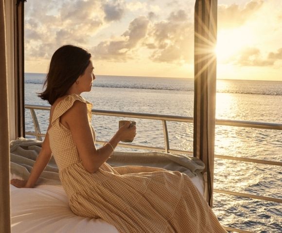 A woman in a light-colored gingham dress holds a mug while sitting near a window overlooking the ocean at sunset.