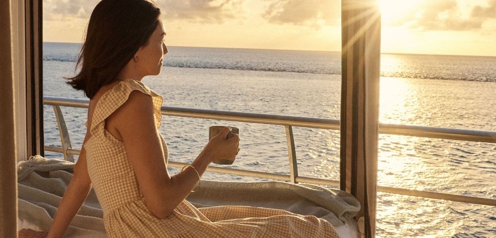 A woman in a light-colored gingham dress holds a mug while sitting near a window overlooking the ocean at sunset.