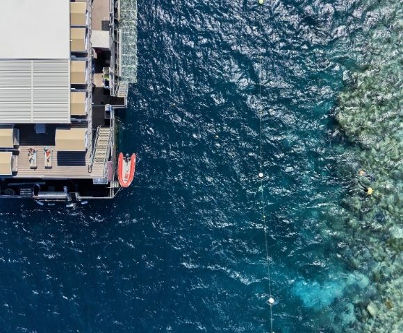 Aerial view of a floating pontoon at a coral reef with people sunbathing on deck and snorkeling in the clear blue water.