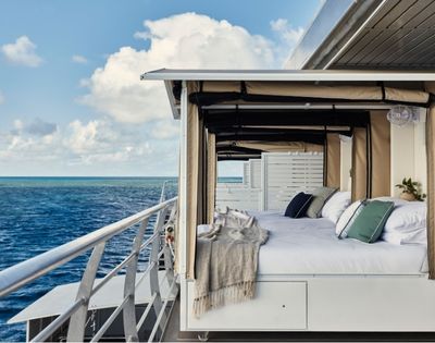 An outdoor bed with white linens and decorative pillows sits on the deck of a boat, overlooking a vast blue ocean under a cloudy sky.