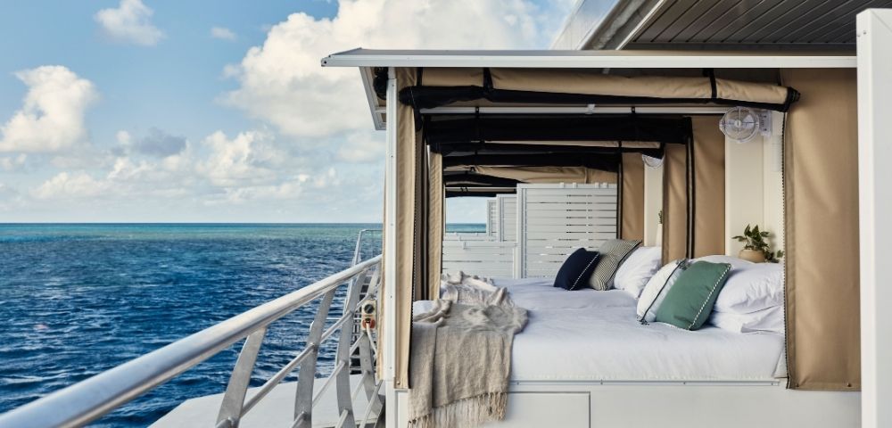 An outdoor bed with white linens and decorative pillows sits on the deck of a boat, overlooking a vast blue ocean under a cloudy sky.