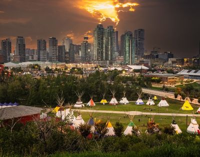 A city skyline at sunset looms over a park featuring a collection of traditional tipis arranged on a grassy field with trees and pathways below.