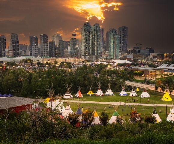 A city skyline at sunset looms over a park featuring a collection of traditional tipis arranged on a grassy field with trees and pathways below.