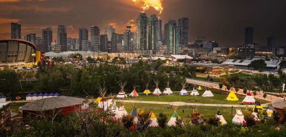 A city skyline at sunset looms over a park featuring a collection of traditional tipis arranged on a grassy field with trees and pathways below.