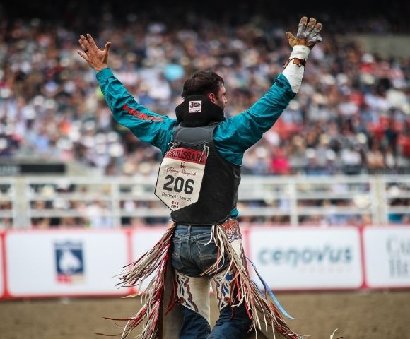A bull rider in a teal shirt and protective vest with number 206 stands in a rodeo arena, arms raised toward a blurred crowd in the background.