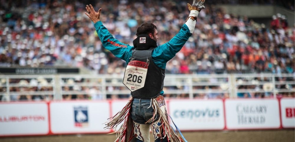 A bull rider in a teal shirt and protective vest with number 206 stands in a rodeo arena, arms raised toward a blurred crowd in the background.
