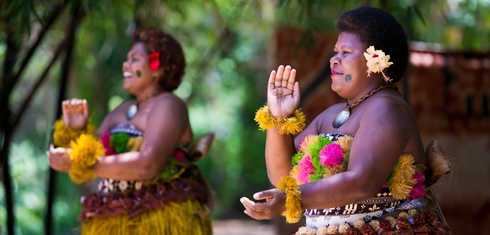 Two women in colorful traditional dress perform a graceful dance in a lush tropical setting, wearing flower garlands and raising their hands.