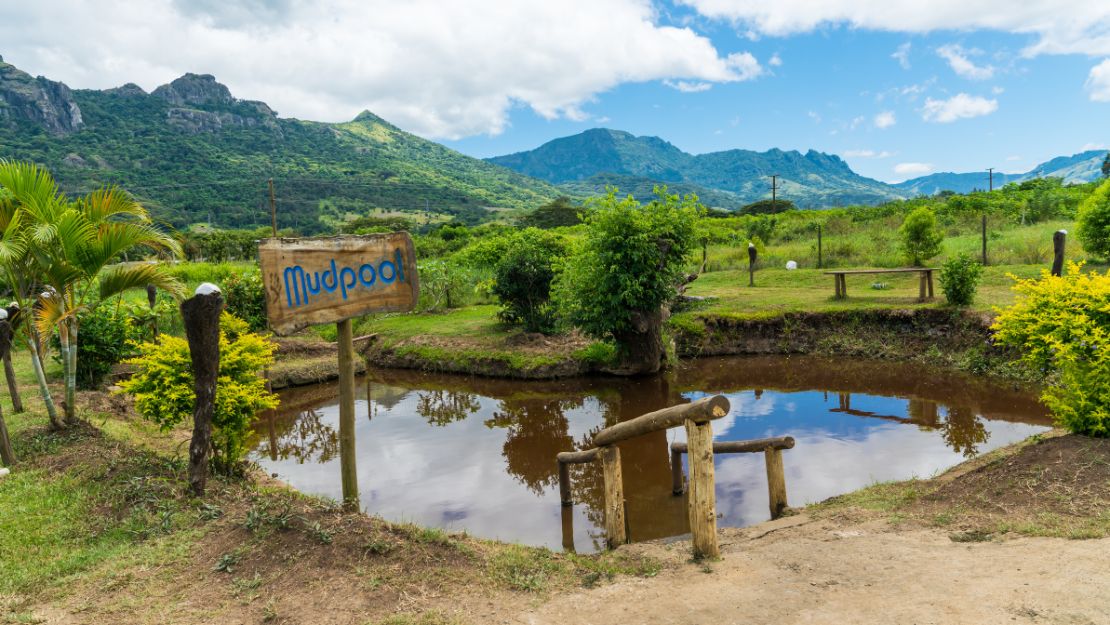 Tropical mountain landscape with a mudpool, rustic footbridge, lush greenery, and a hand-painted “Mudpool” sign beneath bright clouds.