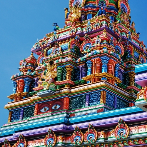 Close-up of a vibrant Hindu temple gopuram, richly covered with colorful deities, guardian faces, ornate carvings, and tiers against blue sky.