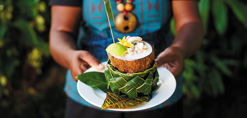 A person in a blue patterned shirt holds a Fijian kokoda dish garnished with a flower.