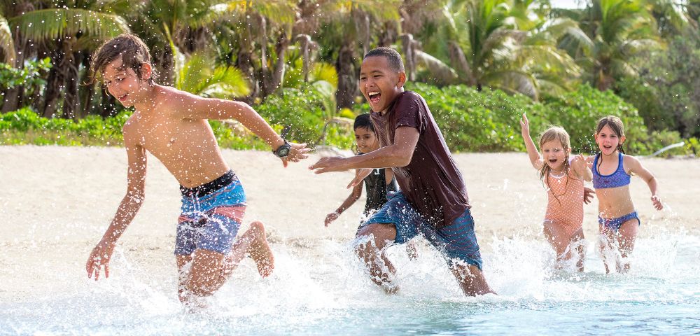 Children laugh and splash through shallow tropical beach water as palm trees sway behind them, capturing a bright, carefree summer moment.