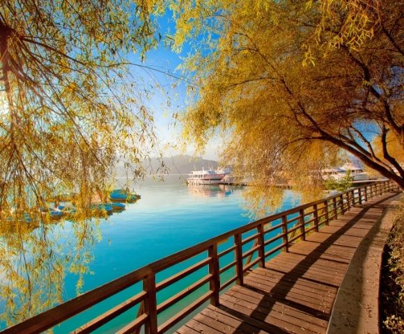 A wooden boardwalk curves along a bright blue lake, framed by golden autumn willow trees with a distant boat docked under a clear sunny sky.
