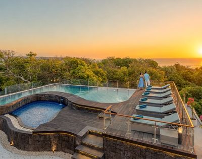 A couple admires a serene sunset over the ocean from the deck of a luxurious infinity pool with chaise lounges.