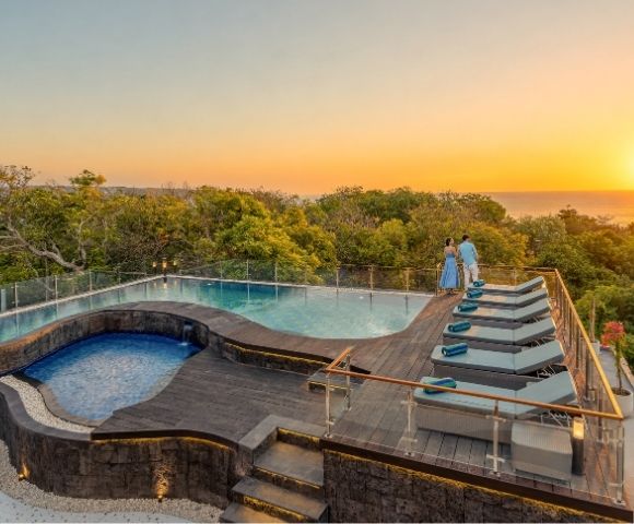 A couple admires a serene sunset over the ocean from the deck of a luxurious infinity pool with chaise lounges.