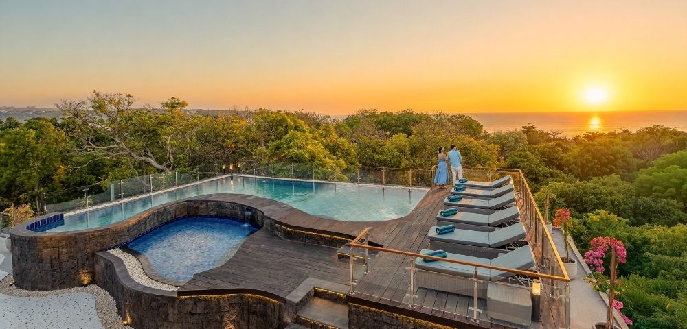 A couple admires a serene sunset over the ocean from the deck of a luxurious infinity pool with chaise lounges.