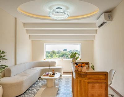 A modern reception area with a curved, cream-colored sectional sofa and a white, organic-shaped coffee table. A wooden reception desk with rattan accents is situated to the right, adorned with decorative elements. A large window overlooks a lush green landscape.