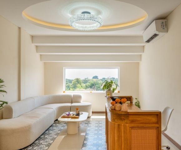A modern reception area with a curved, cream-colored sectional sofa and a white, organic-shaped coffee table. A wooden reception desk with rattan accents is situated to the right, adorned with decorative elements. A large window overlooks a lush green landscape.