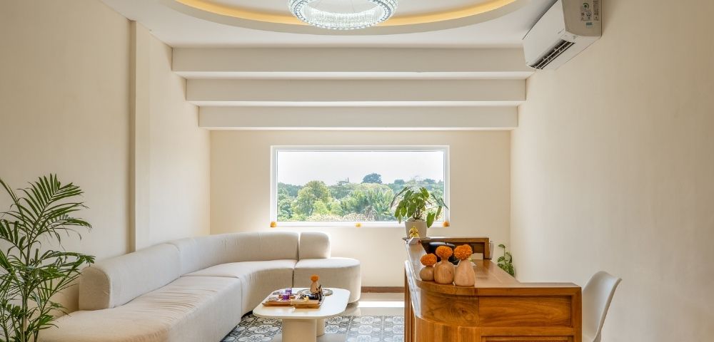 A modern reception area with a curved, cream-colored sectional sofa and a white, organic-shaped coffee table. A wooden reception desk with rattan accents is situated to the right, adorned with decorative elements. A large window overlooks a lush green landscape.