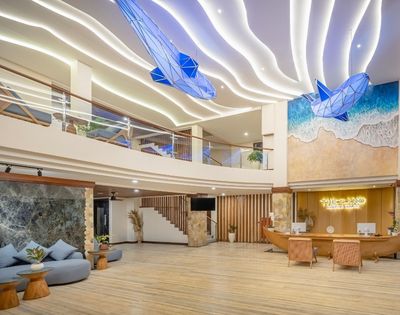 A modern hotel lobby with a spacious, multi-level design featuring wavy illuminated ceilings and two large blue whale sculptures suspended overhead. A beach mural decorates the wall behind a reception desk shaped like a boat.