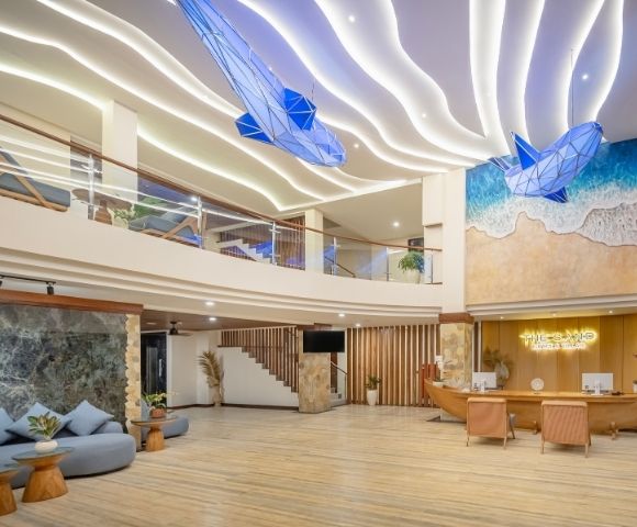 A modern hotel lobby with a spacious, multi-level design featuring wavy illuminated ceilings and two large blue whale sculptures suspended overhead. A beach mural decorates the wall behind a reception desk shaped like a boat.
