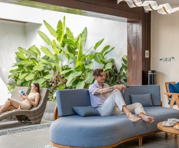 A man with tattoos on his arms sits on a blue couch, holding a remote control. A woman reclines on a brown chaise lounge, reading a book. Lush green plants fill the background.