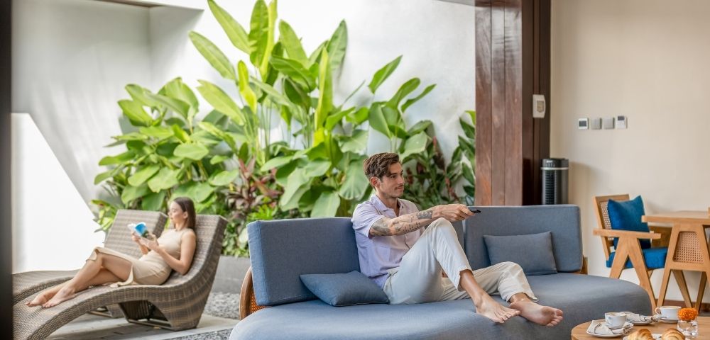 A man with tattoos on his arms sits on a blue couch, holding a remote control. A woman reclines on a brown chaise lounge, reading a book. Lush green plants fill the background.