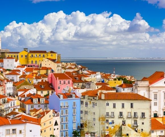 Colorful Lisbon hillside rooftops overlooking the blue Tagus River, with pastel buildings, red tiles, and dramatic white clouds under a sunny sky.