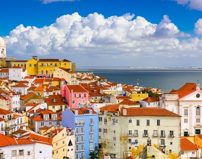 Colorful Lisbon hillside rooftops overlooking the blue Tagus River, with pastel buildings, red tiles, and dramatic white clouds under a sunny sky.