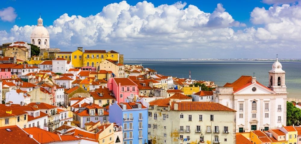 Colorful Lisbon hillside rooftops overlooking the blue Tagus River, with pastel buildings, red tiles, and dramatic white clouds under a sunny sky.