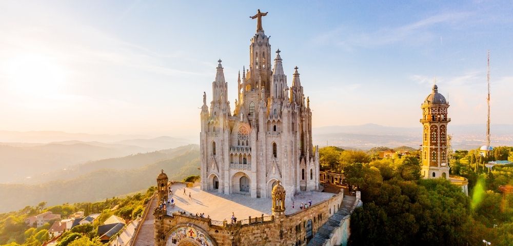 Aerial view of Barcelona’s ornate Sacred Heart church atop Tibidabo, crowned by a cross and glowing in warm sunset above wooded hills.