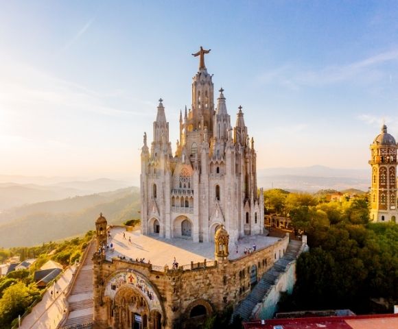 Aerial view of Barcelona’s ornate Sacred Heart church atop Tibidabo, crowned by a cross and glowing in warm sunset above wooded hills.