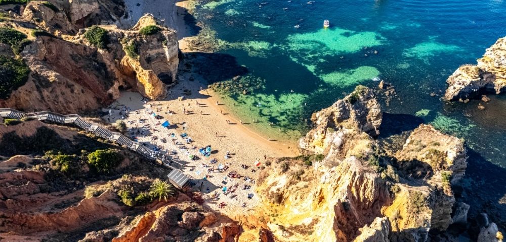 Aerial view of a secluded Algarve beach, with turquoise water, golden cliffs, rocky sea stacks, swimmers and sunbathers creating a serene scene.