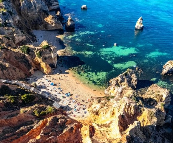 Aerial view of a secluded Algarve beach, with turquoise water, golden cliffs, rocky sea stacks, swimmers and sunbathers creating a serene scene.Aerial view of a secluded Algarve beach, with turquoise water, golden cliffs, rocky sea stacks, swimmers and sunbathers creating a serene scene.