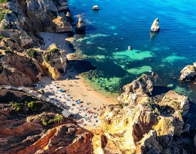 Aerial view of a secluded Algarve beach, with turquoise water, golden cliffs, rocky sea stacks, swimmers and sunbathers creating a serene scene.