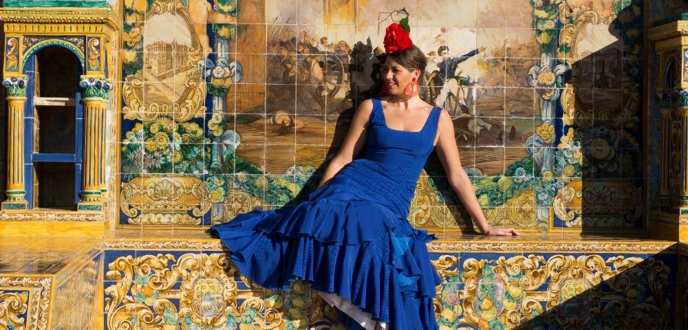 Woman in a flowing blue dress and red flower sits before an ornate ceramic mural labeled “Madrid,” evoking elegance and cultural charm.