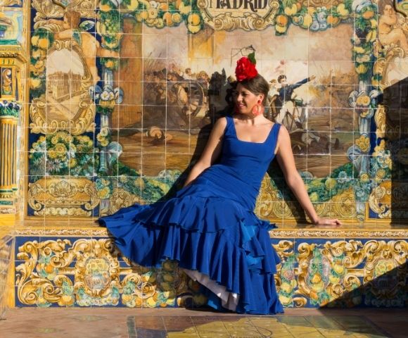 Woman in a flowing blue dress and red flower sits before an ornate ceramic mural labeled “Madrid,” evoking elegance and cultural charm.