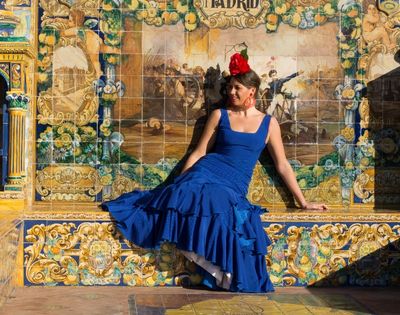 Woman in a flowing blue dress and red flower sits before an ornate ceramic mural labeled “Madrid,” evoking elegance and cultural charm.