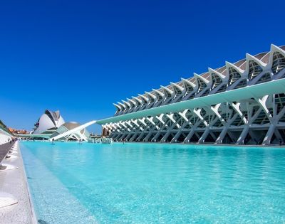 A modern architectural complex featuring a large pool of clear blue water, with a striking building design and a cloudless blue sky in the background.