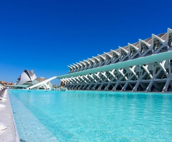 A modern architectural complex featuring a large pool of clear blue water, with a striking building design and a cloudless blue sky in the background.