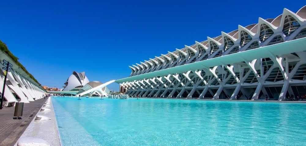 A modern architectural complex featuring a large pool of clear blue water, with a striking building design and a cloudless blue sky in the background.